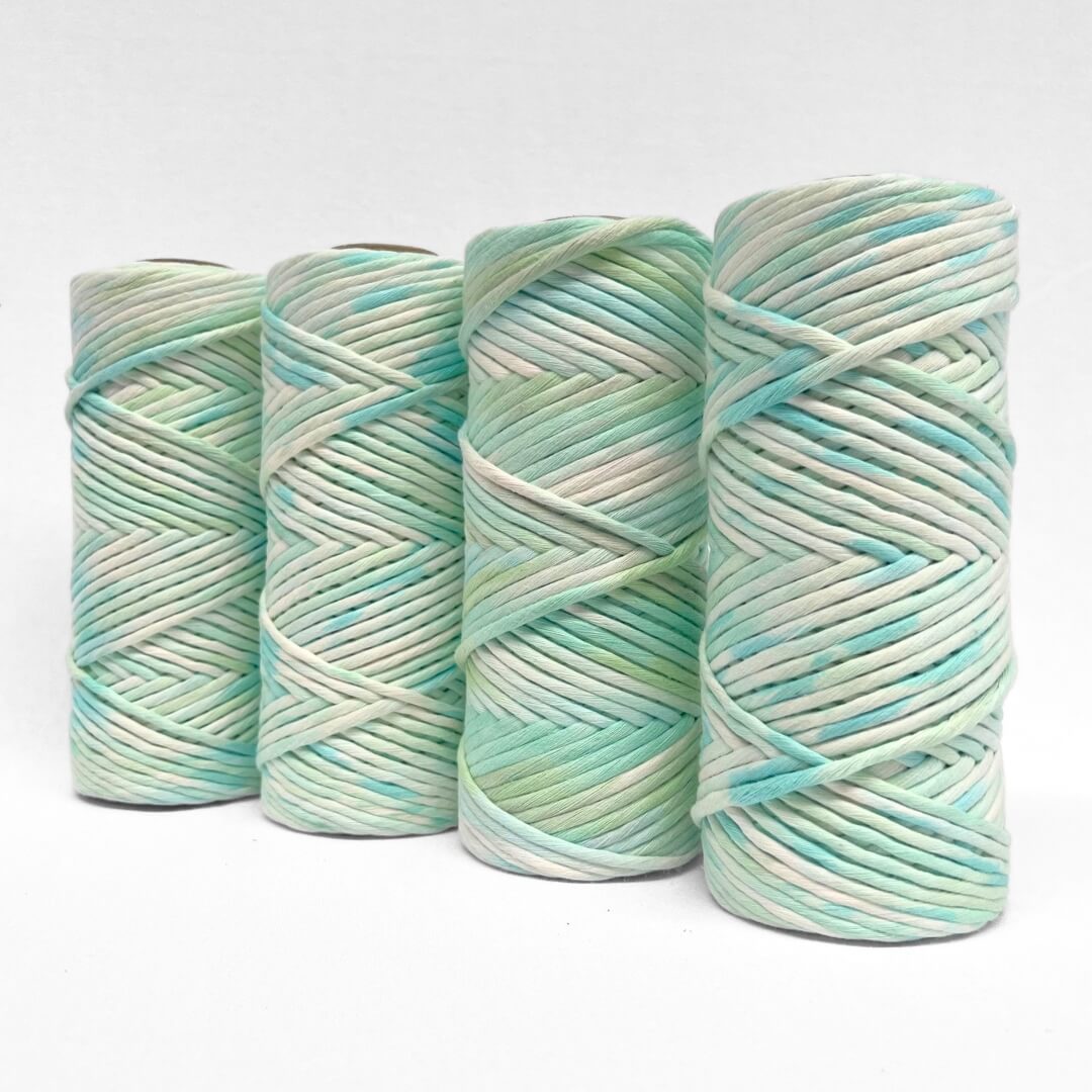 four rolls of blue and green hand painted macrame string standing upright on white background 