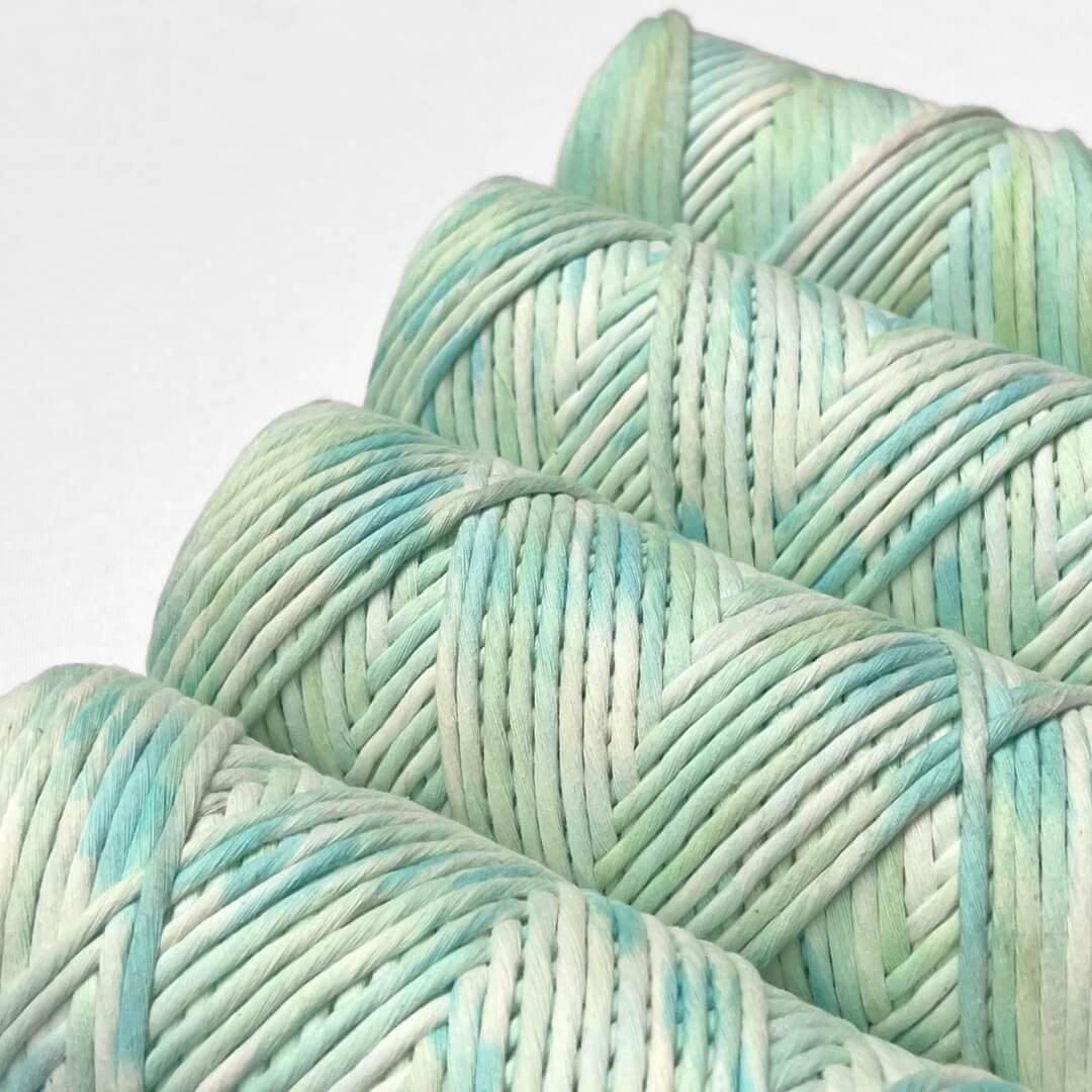 four rolls of blue and green hand painted macrame string standing upright on white background 