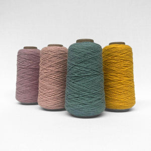 four rolls of wool thin string in teal yellow blush and purple standing on angle standing upright on white wall